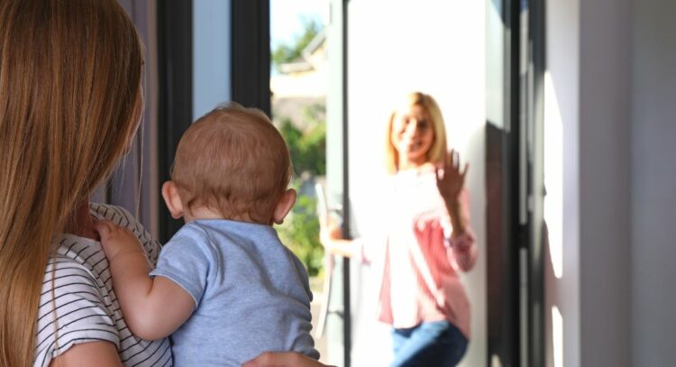 une femme sort par la porte d'entree en saluant une jeune femme qui porte un bebe dans ses bras